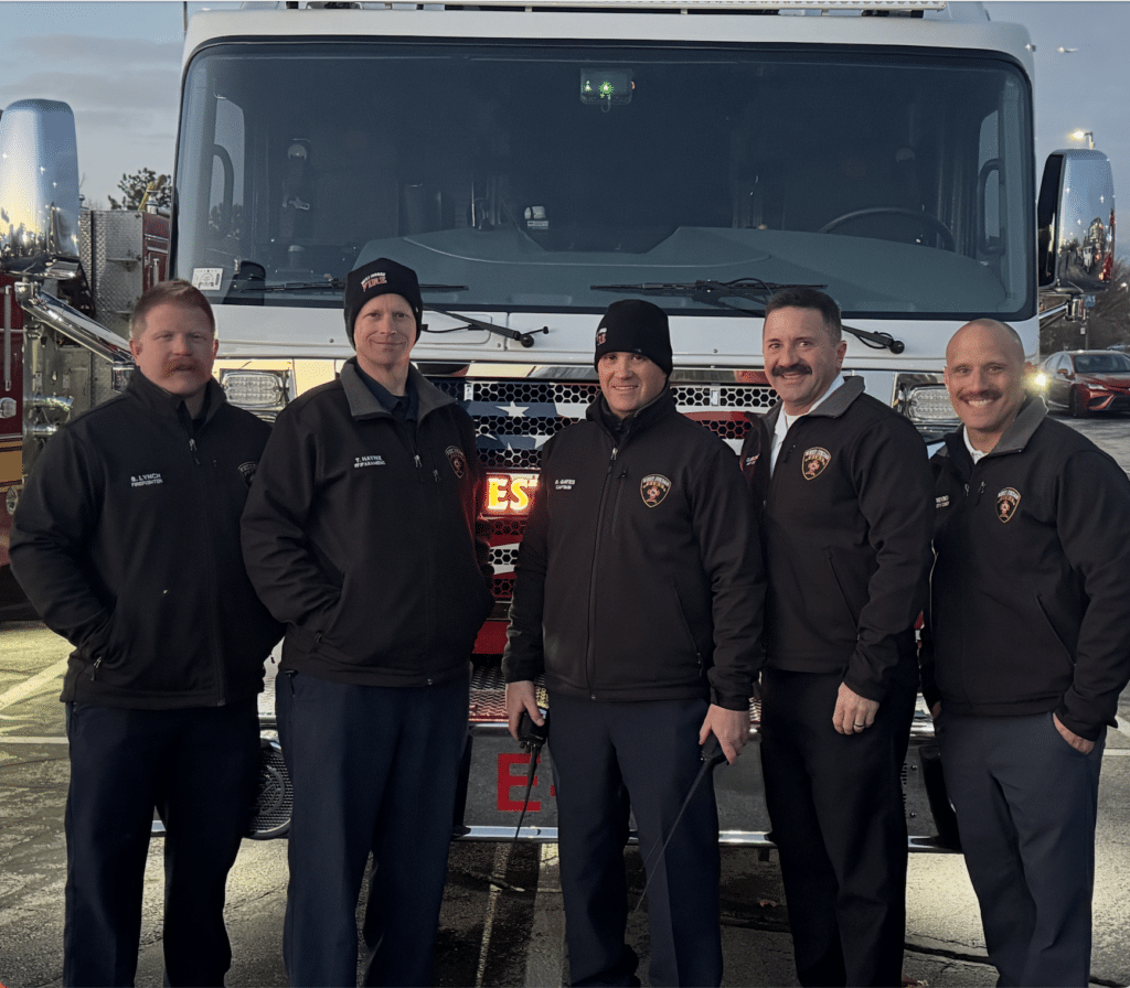 Five firefighters standing in front of a fire truck, all wearing black jackets and hats with fire department emblems. They are smiling, and its early evening as the sky is dimly lit.